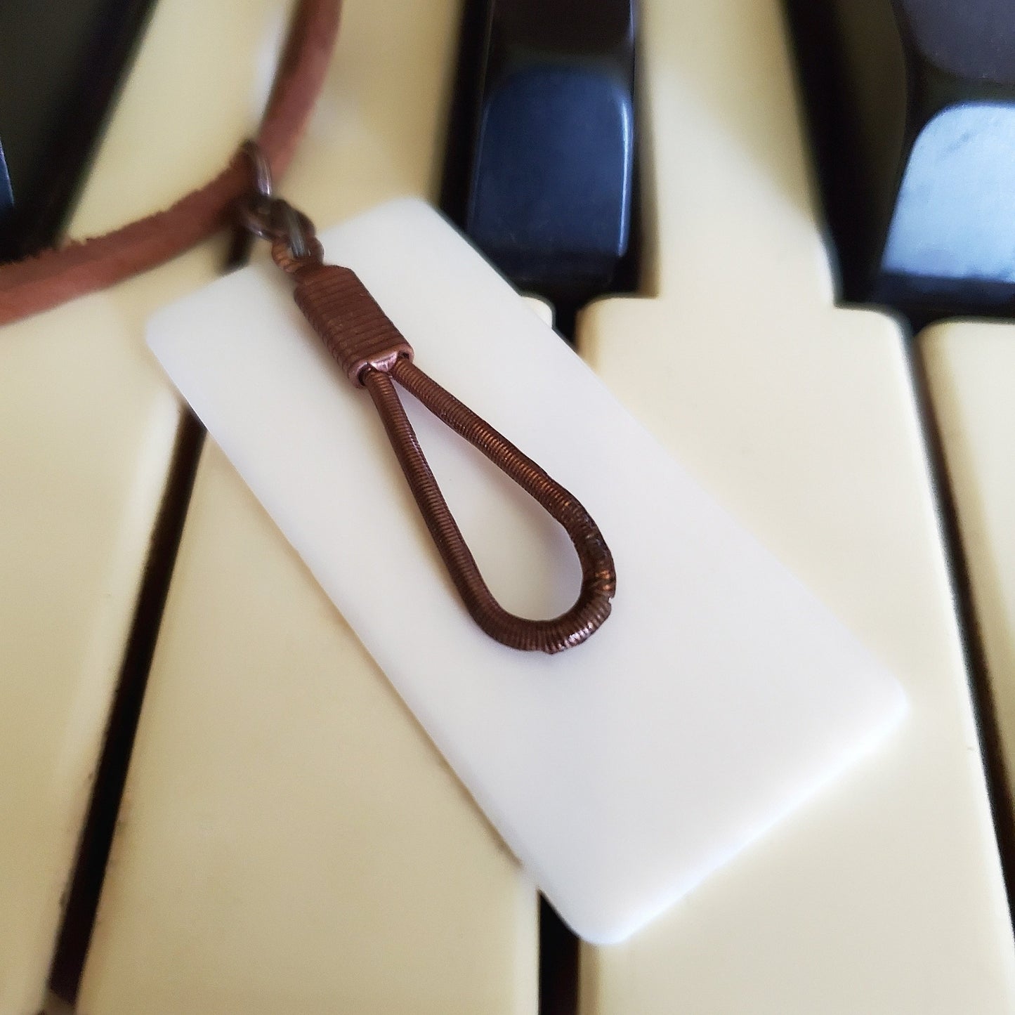 close-up of a necklace made from an upcycled ivory piano key topper and a teardrop shaped upcycled piano string both hanging on a brown suede chord - necklace is on piano keys
