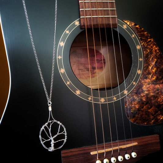 a necklace made with a chain on which hangs a pendant in the shape of a tree with a circle around it - the tree is made of silver coloured wire and the circle is made from upcycled guitar strings - the necklace hangs in front of a black guitar