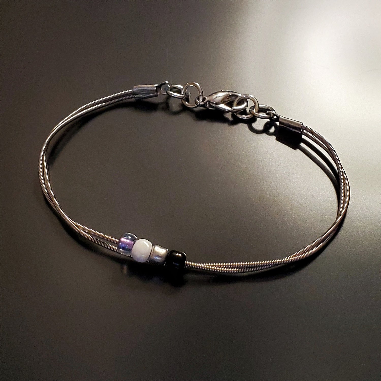 silver coloures clasp style bracelet with 4 glass beads representing the asexual pride flag (black, grey, white & purple)