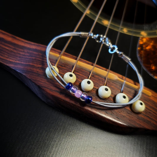 guitar string bracelet with blue, purple and pink bead representing the bisexual pride flag lying on a black guitar