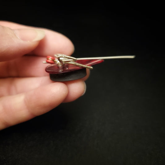 fingers holding a magnet made from an upcycled guitar string shaped like a dragonfly sitting on a dark red guitar pick - black background