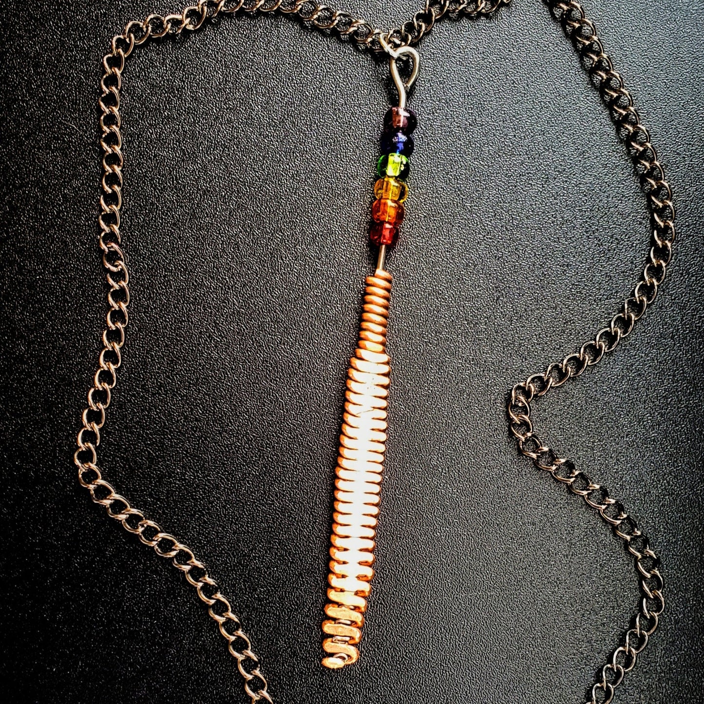 Hammered piano string pendant with 6 LGBTQ flag coloured beads with chain on a piano black background