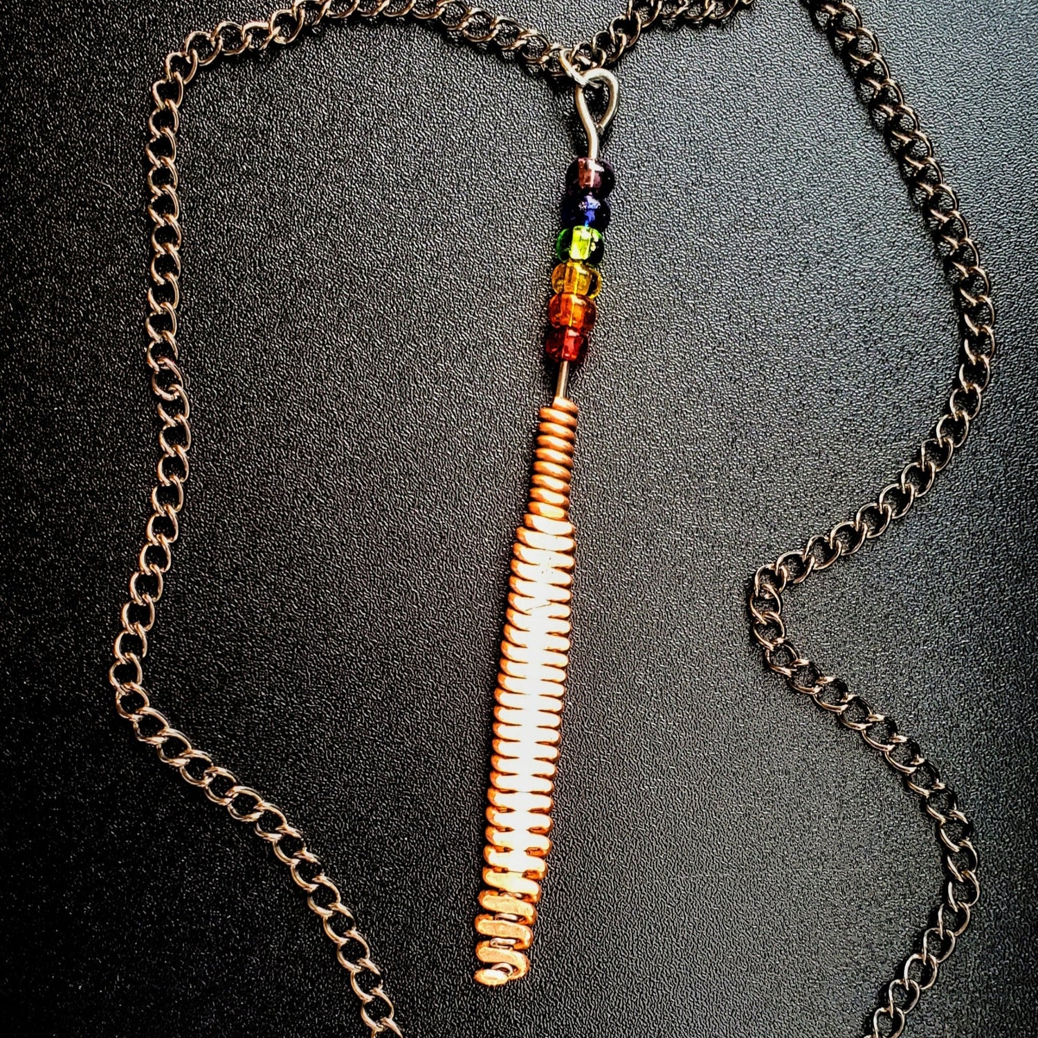 Hammered piano string pendant with 6 LGBTQ flag coloured beads with chain on a piano black background