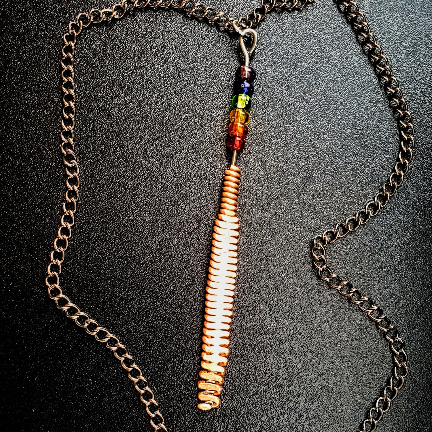 Hammered piano string pendant with 6 LGBTQ flag coloured beads with chain on a piano black background