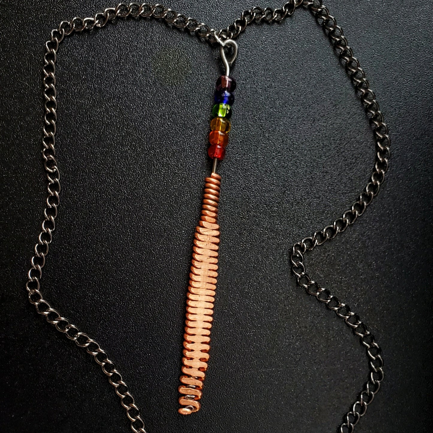 Hammered piano string pendant with 6 LGBTQ flag coloured beads with chain on a piano black background