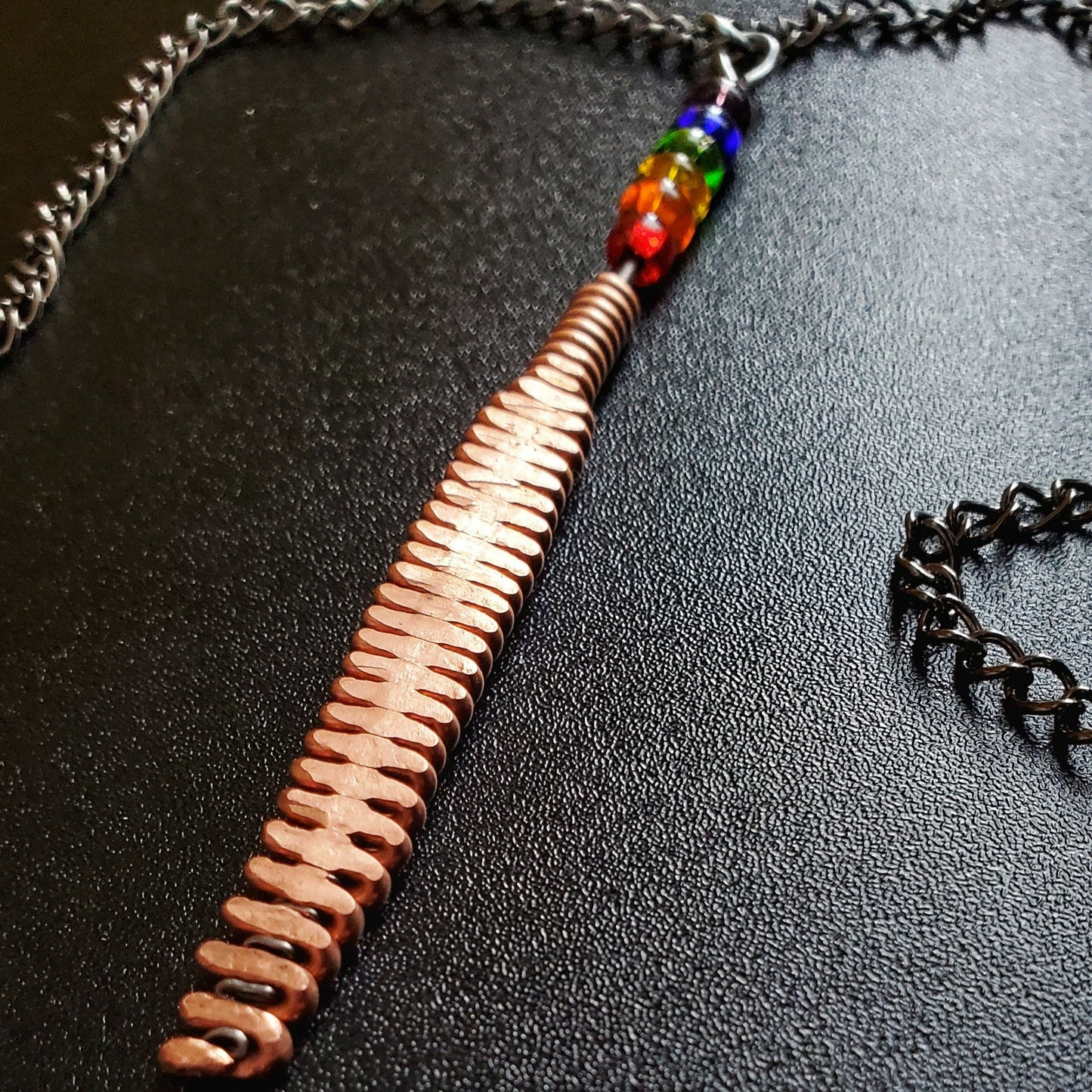 Hammered piano string pendant with 6 LGBTQ flag coloured beads with chain on a piano black background