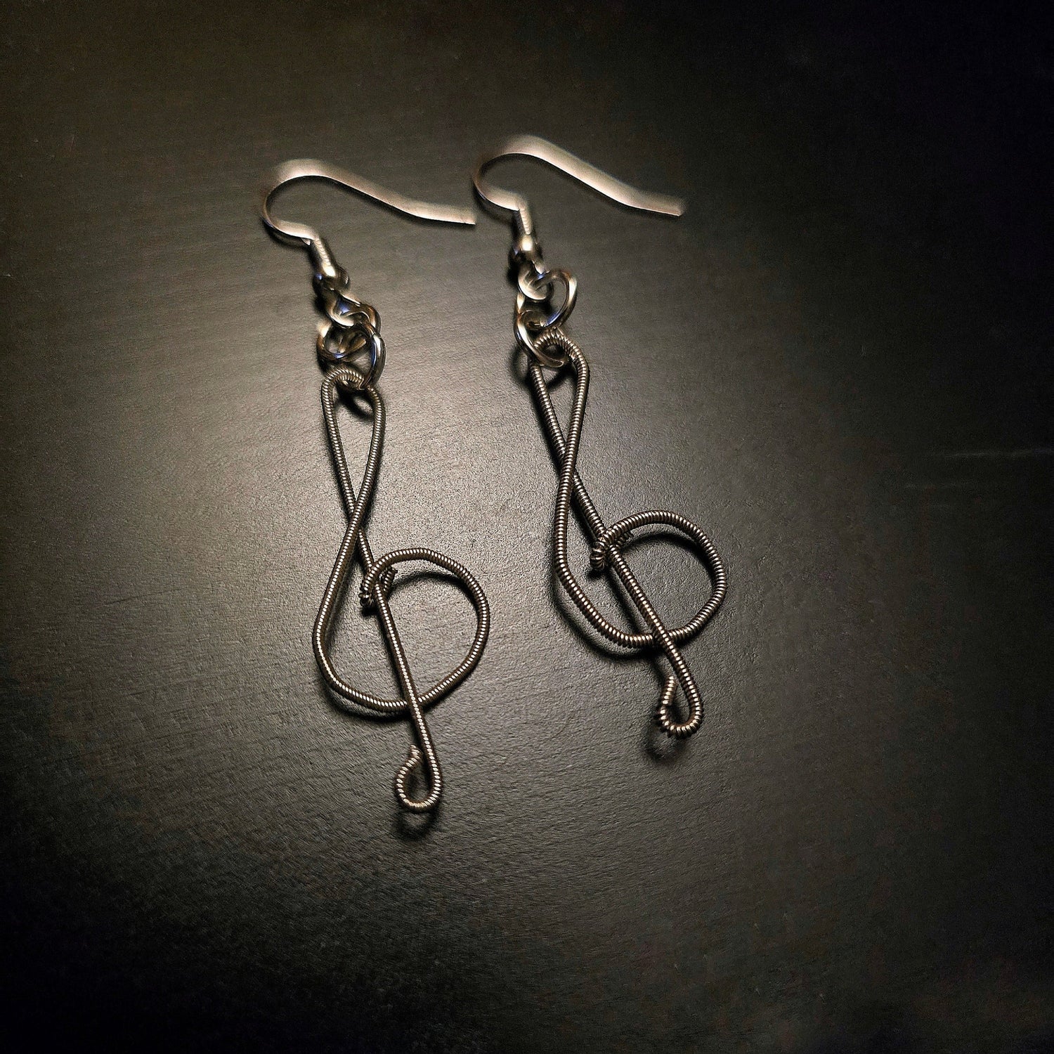 pair of earrings in the shape of treble clefs, made from upcycled guitar strings lying a black background