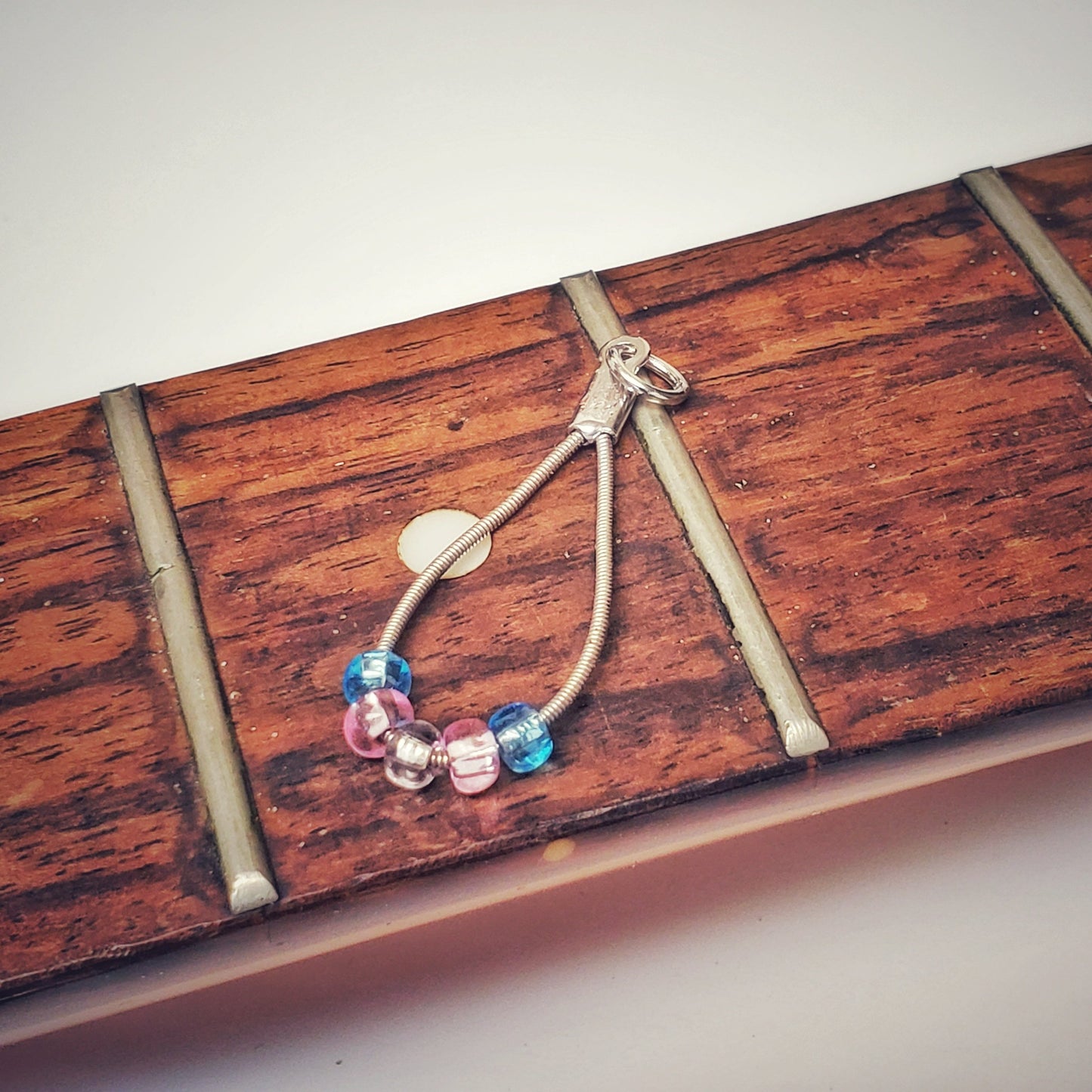 silver coloured teardrop pendant made from upcycled guitar strings - there are 5 beads representing the colours of the transgender pride flag - 2 blue, 2 pink and 1 white - pendant is sitting on the neck of a guitar which has no stings