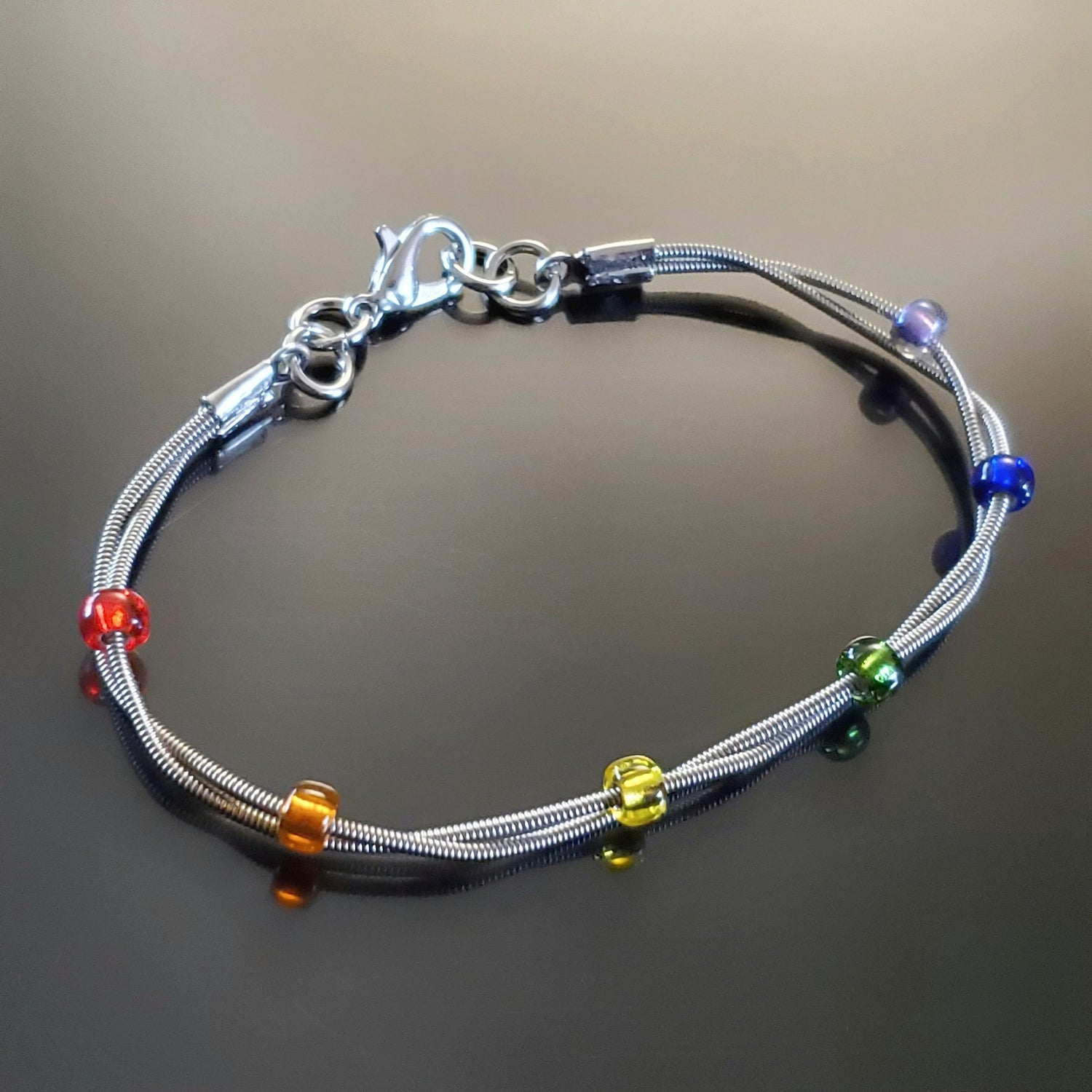 clasp style bracelet made from two upcycled guitar strings - 6 glass beads represent the colours of the LGBTQ flag (red, orange, yellow, green, blue and purple)