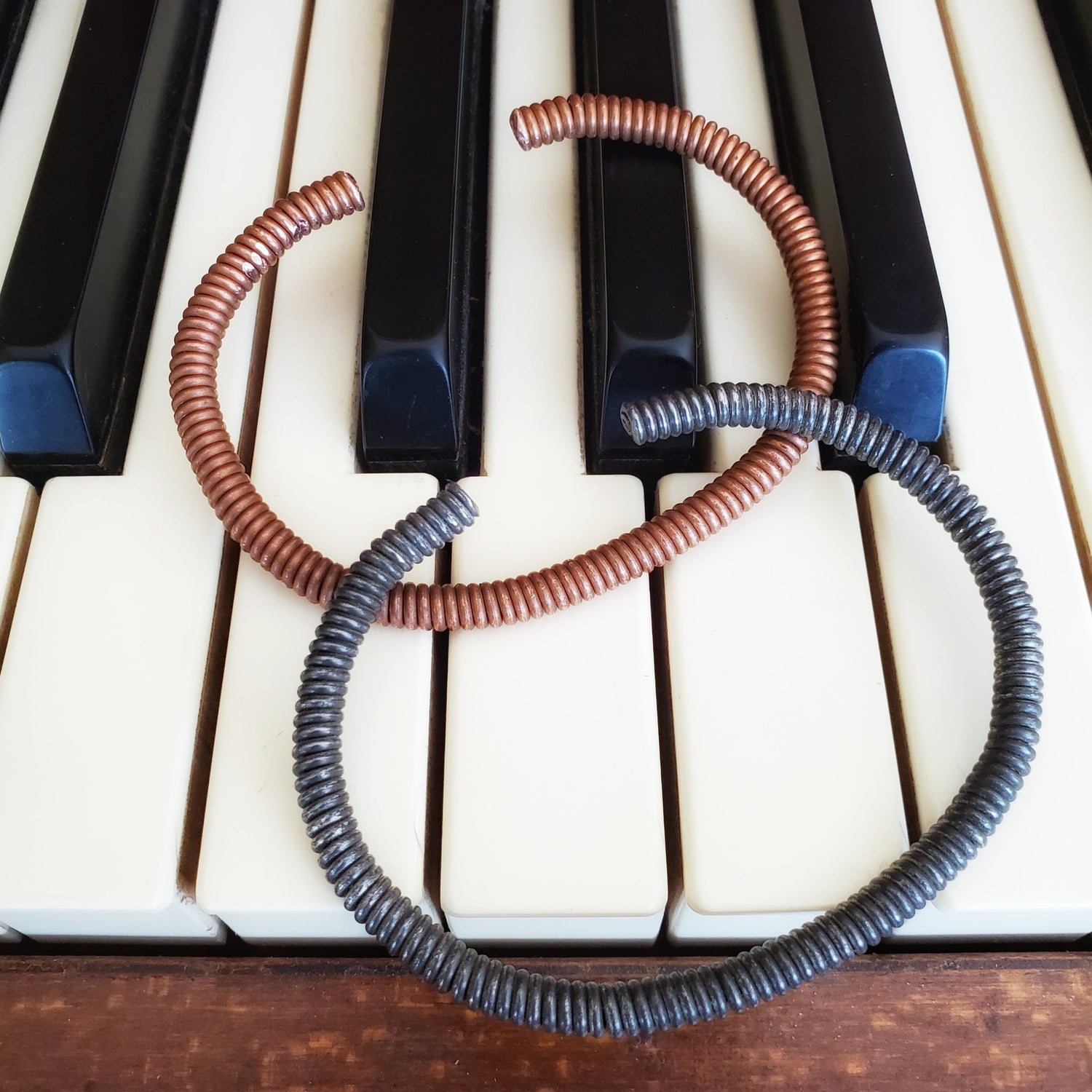 copper and charcoal grey cuff style bracelets made from an upcycled piano strings - both bracelets are lying on piano keys