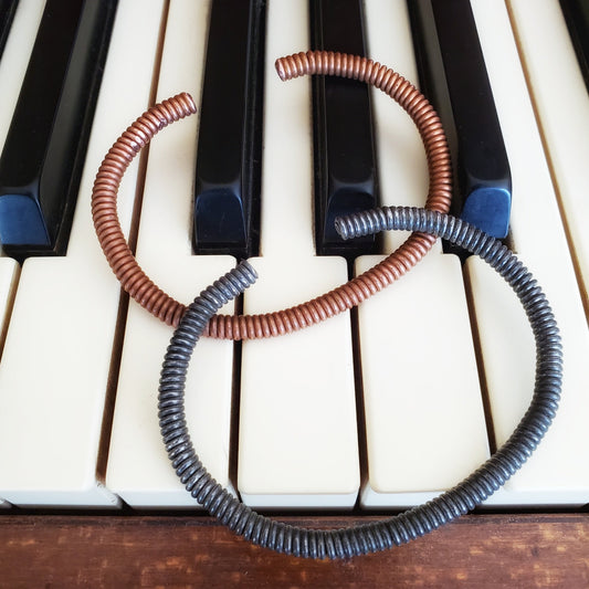 copper and charcoal grey cuff style bracelets made from an upcycled piano strings - both bracelets are lying on piano keys