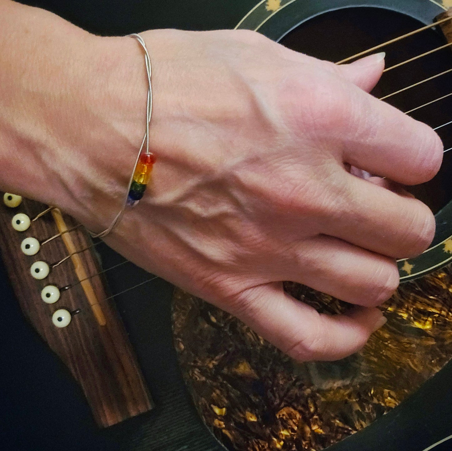 Hand playing a guitar wearing a guita bangle with 6 beads representing the LGBTQ pride flag