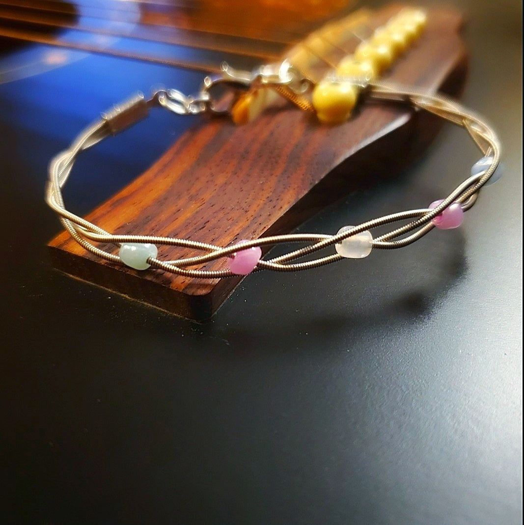 clasp style bracelet made from braided upcycled guitar strings with glass beads representing the Trans Pride flag sitting on the bridge of a black guitar