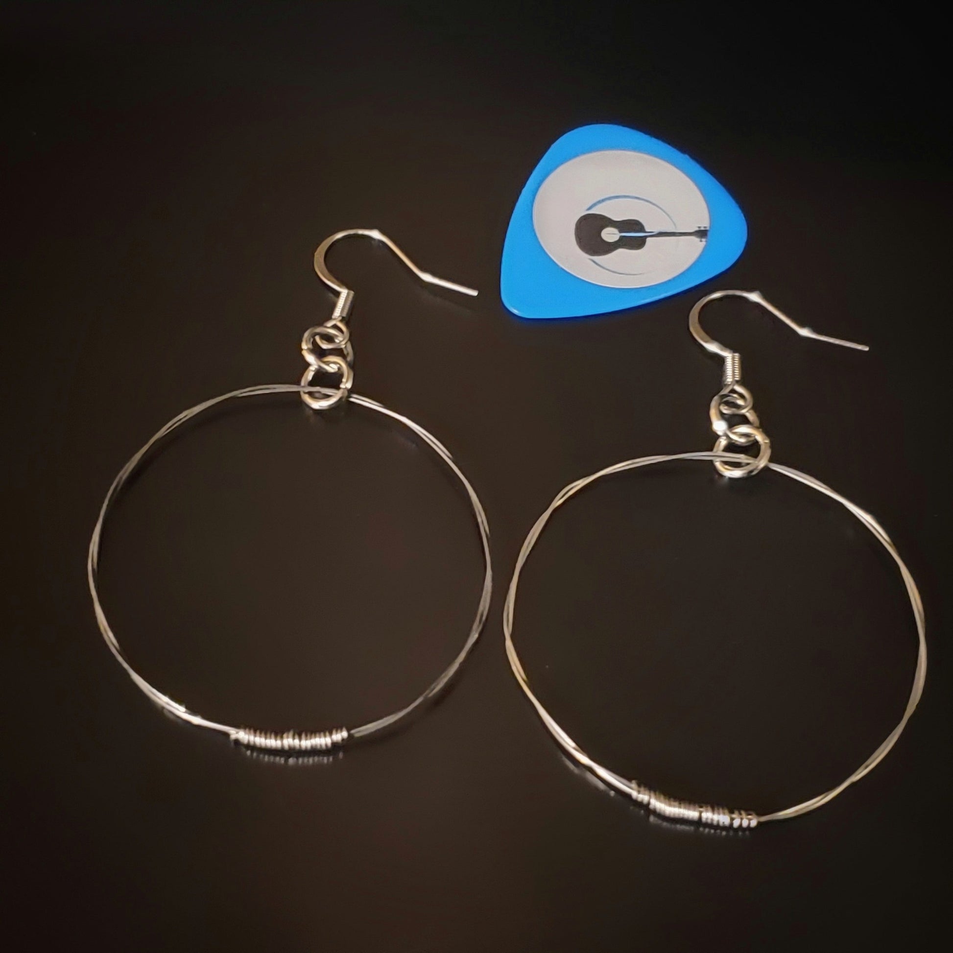 silver coloured hoop style earrings made from upcycled guitar strings - above is a blue guitar pick with a picture of a black guitar- black background