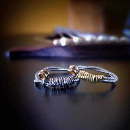 two rings are sitting on a black guitar - both are made from upcycled guitar strings, both are silver but the left one has silver coloured wire and the right one has gold coloured wire