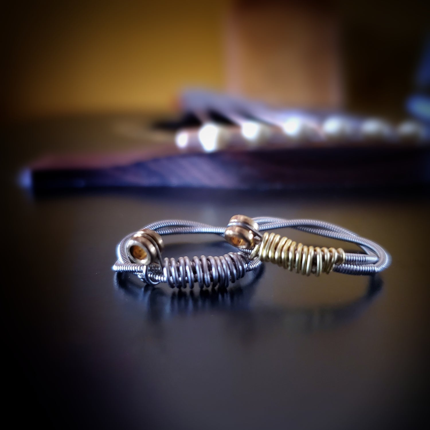 two rings are sitting on a black guitar - both are made from upcycled guitar strings, both are silver but the left one has silver coloured wire and the right one has gold coloured wire