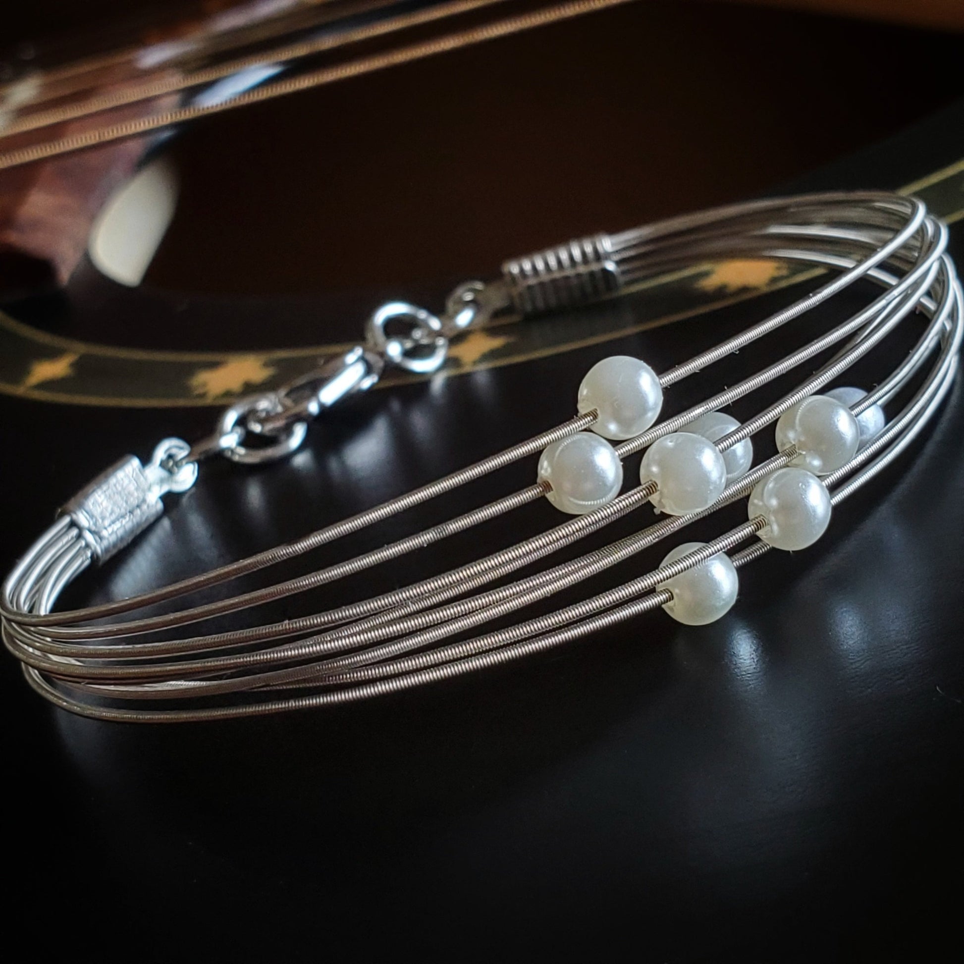 clasp style bracelet made from several strands of upcycled guitar strings on each one is a white glass bead- bracelet is lying on the body of a black guitar