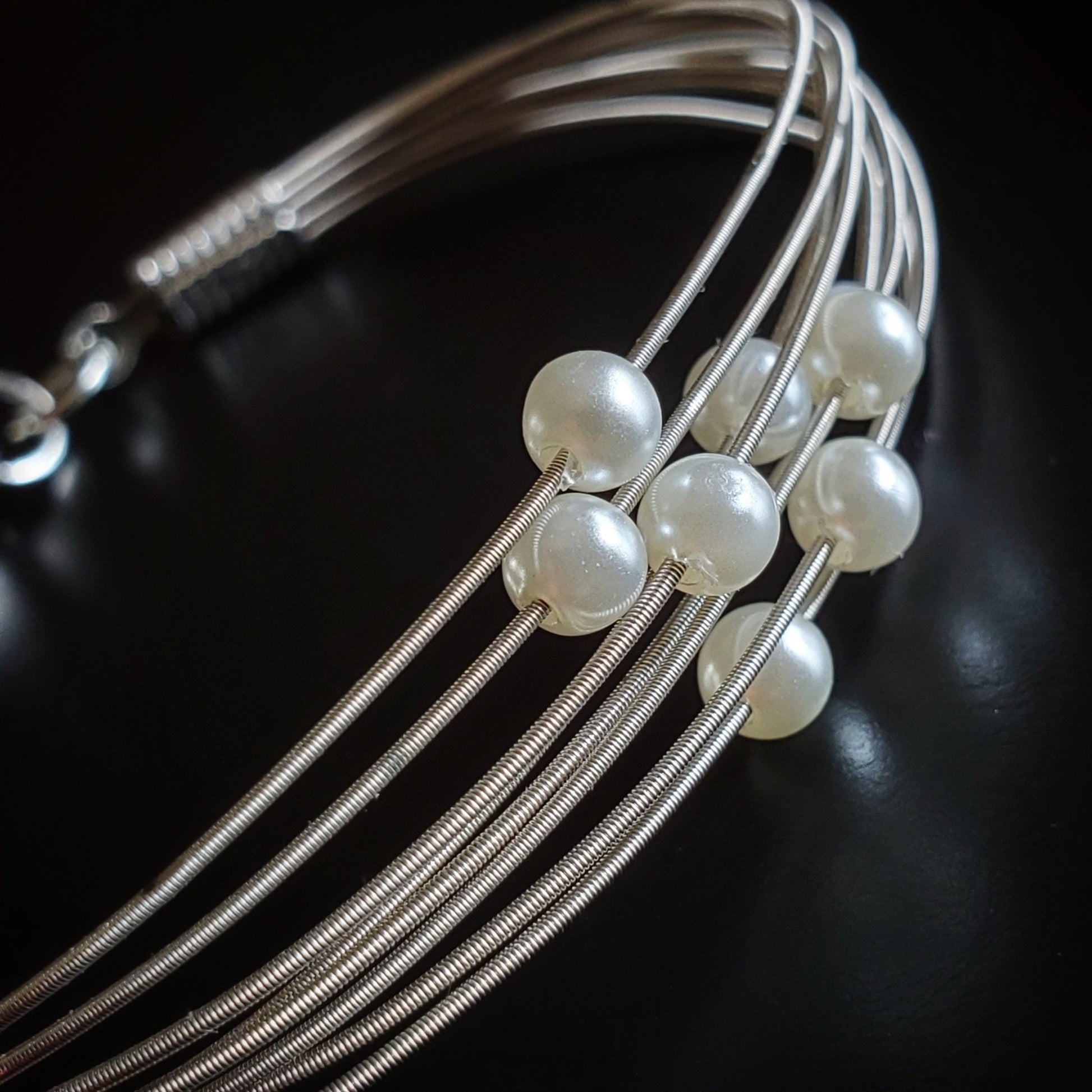 close-up of clasp style bracelet made from several strands of upcycled guitar strings on each one is a white glass bead