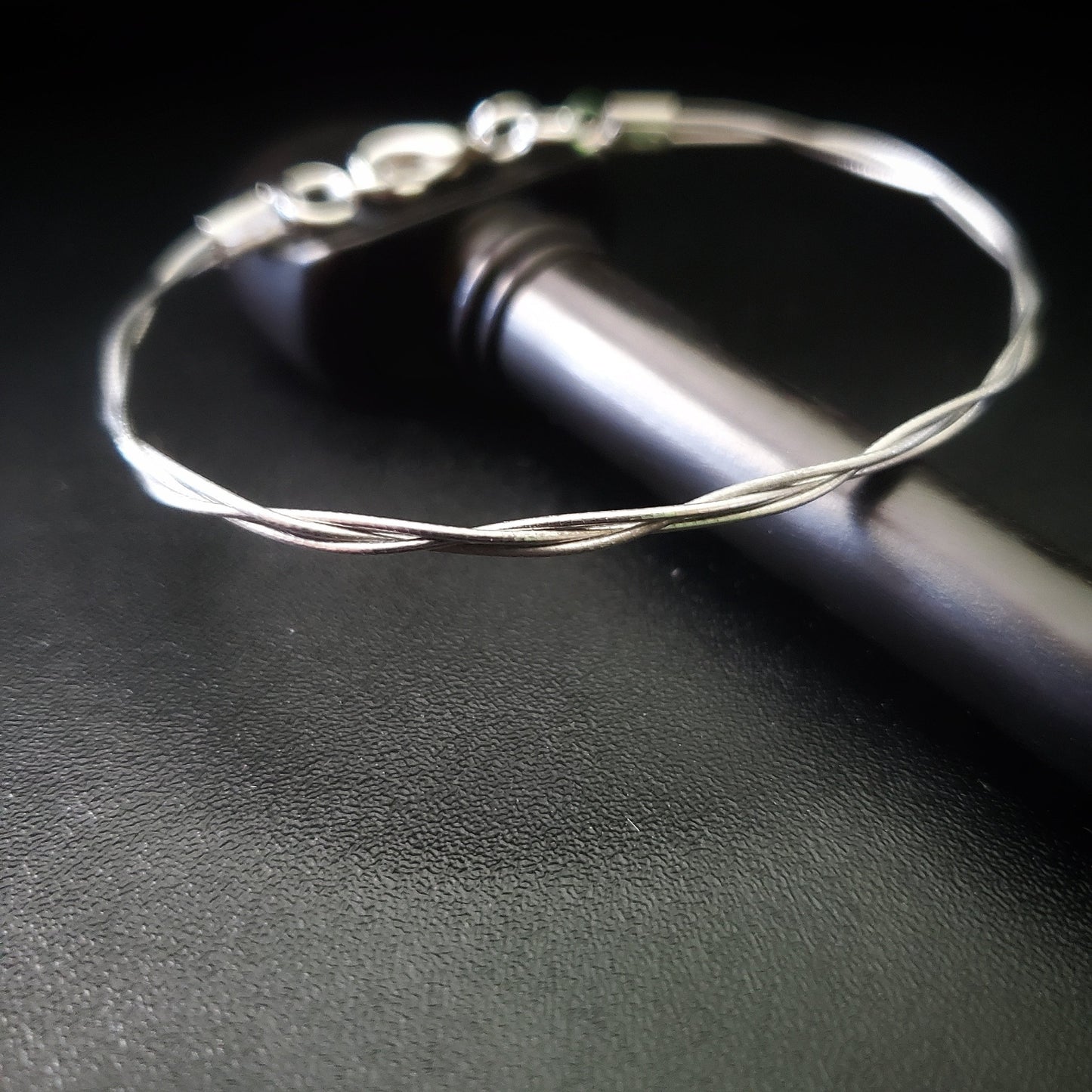 silver coloured bracelet made from 3 cello strings braided together sitting on a black cello tuning peg on a black background
