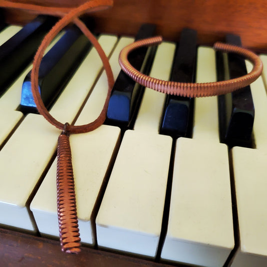 a clasp style bracelet made from an upcycled copper piano string hammered flat on its left is a necklace made from a piece of hammered piano string and a piece of brown suede cord both are sitting on piano keys
