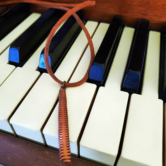 necklace made from a brown suede lanyard on which hangs a pendant made from a copper piano string hammered flat - the necklace rests on a piano keyboard