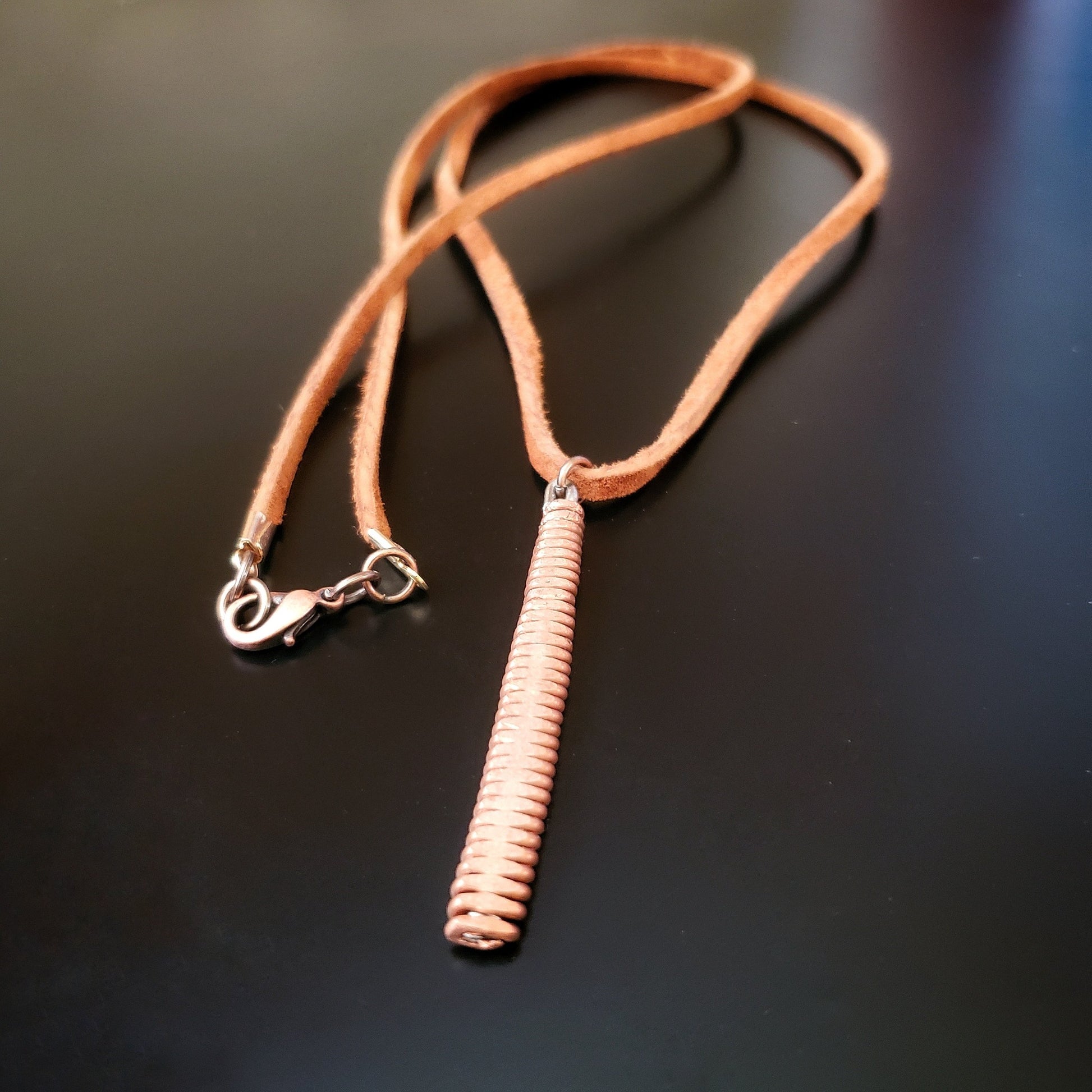 necklace made from a brown suede lanyard on which hangs a pendant made from a copper piano string hammered flat