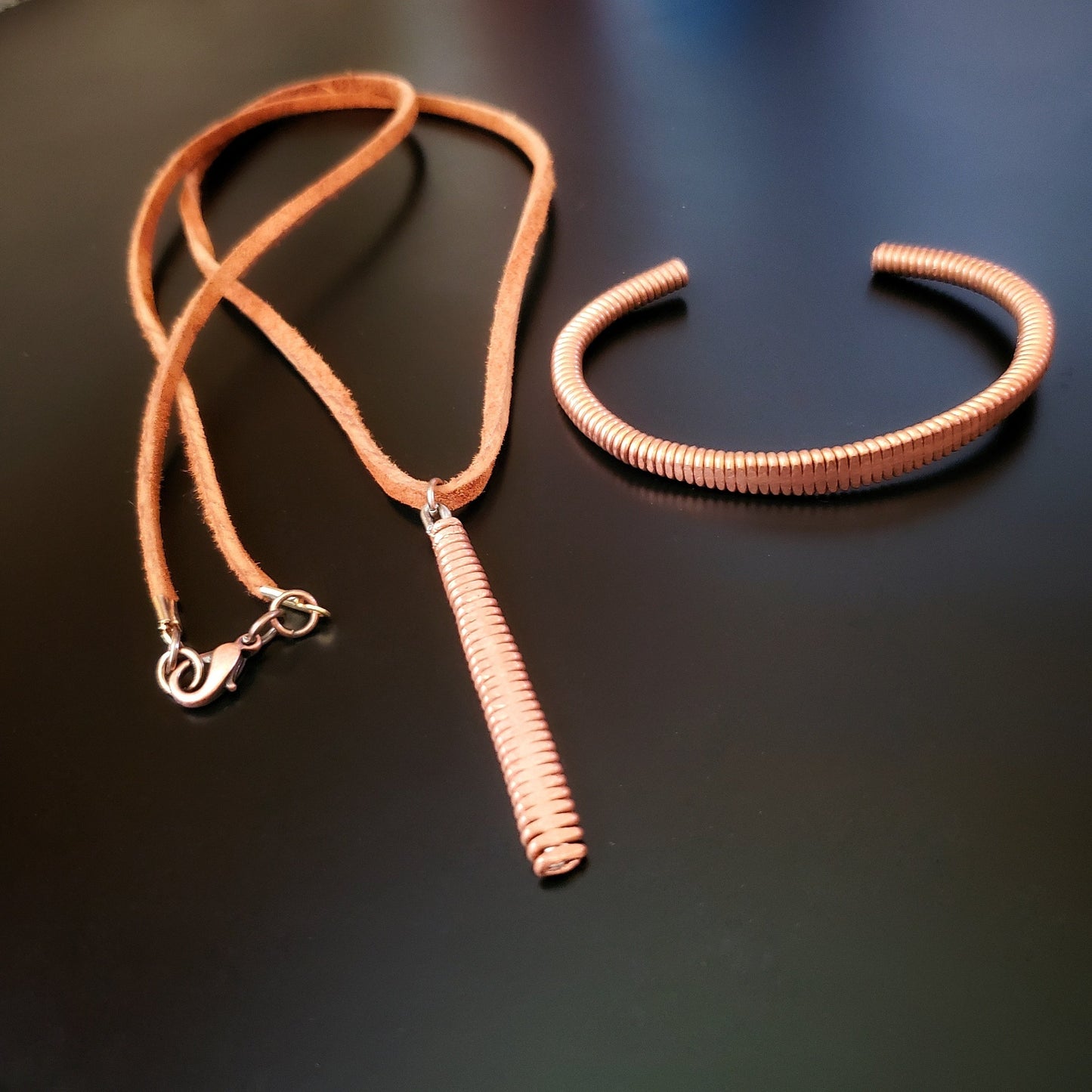 a clasp style bracelet made from an upcycled copper piano string hammered flat on its left is a necklace made from a piece of hammered piano string and a piece of brown suede cord
