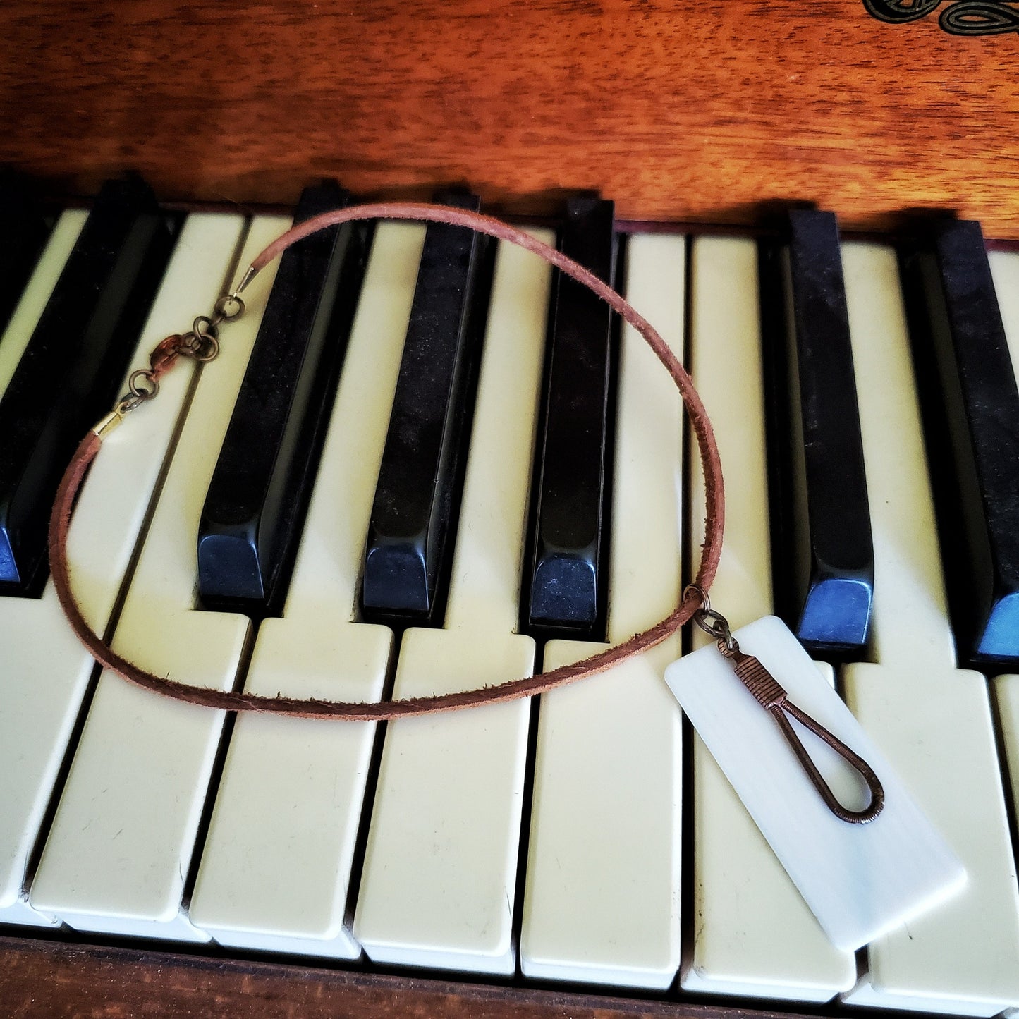 necklace made from an upcycled ivory piano key topper and a teardrop shaped upcycled piano string both hanging on a brown suede chord - necklace is on piano keys