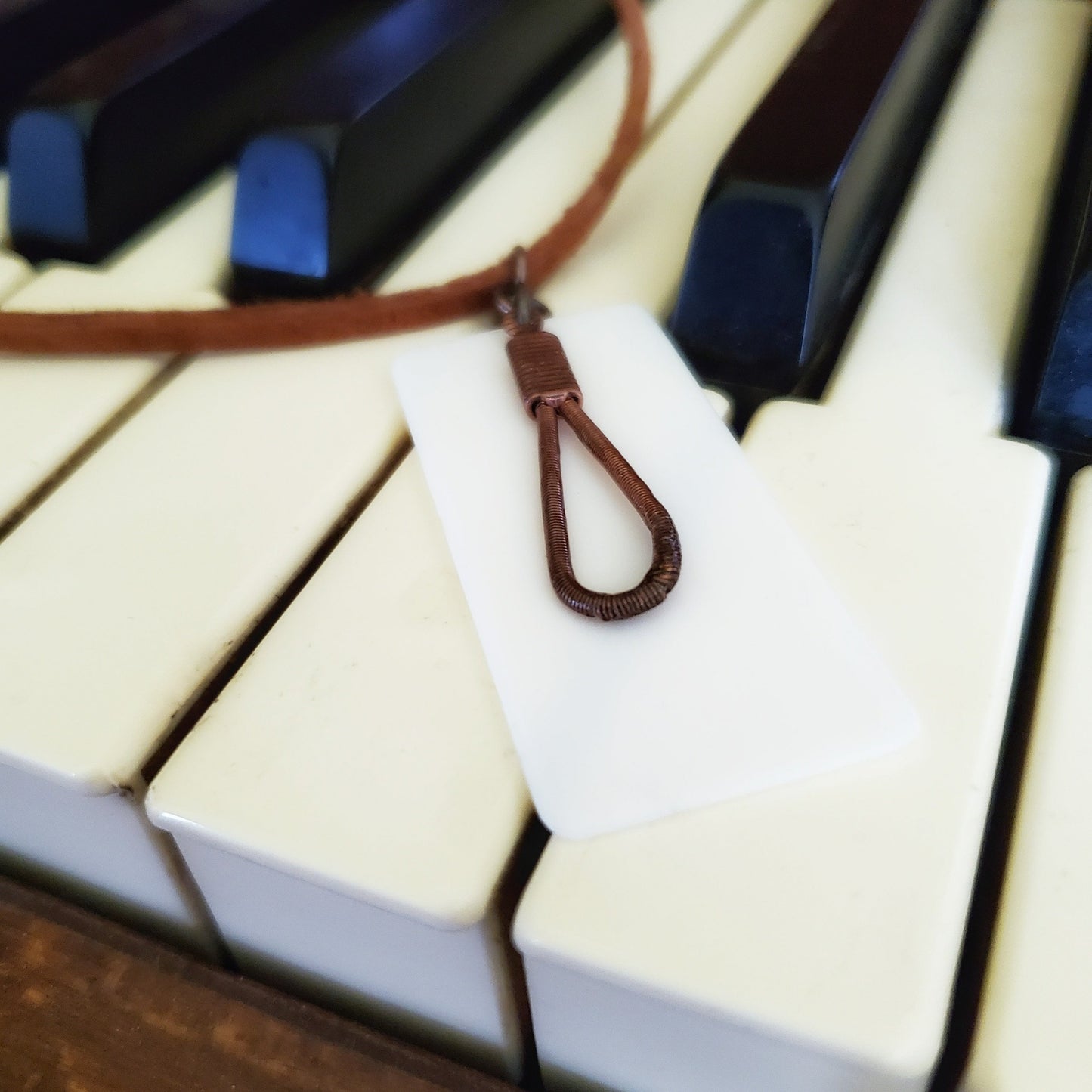 close-up of a necklace made from an upcycled ivory piano key topper and a teardrop shaped upcycled piano string both hanging on a brown suede chord - necklace is on piano keys