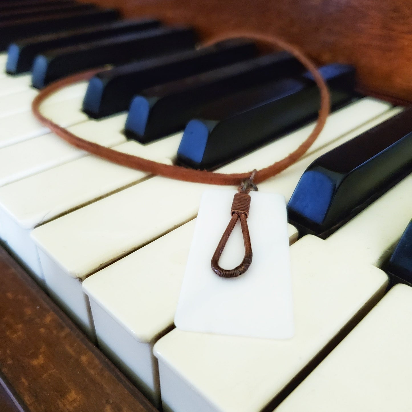 necklace made from an upcycled ivory piano key topper and a teardrop shaped upcycled piano string both hanging on a brown suede chord - necklace is on piano keys
