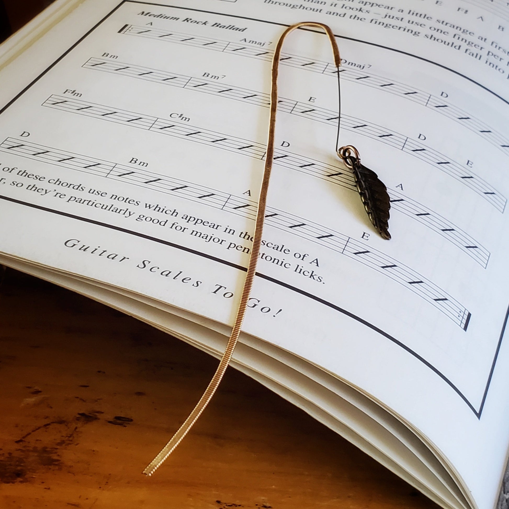 hook style bookmark made from an upcycled guitar string and a leaf shaped charm- background is the page of a book with the words Guitar Scales to Go and some guitar chords for a Medium Rock Ballad