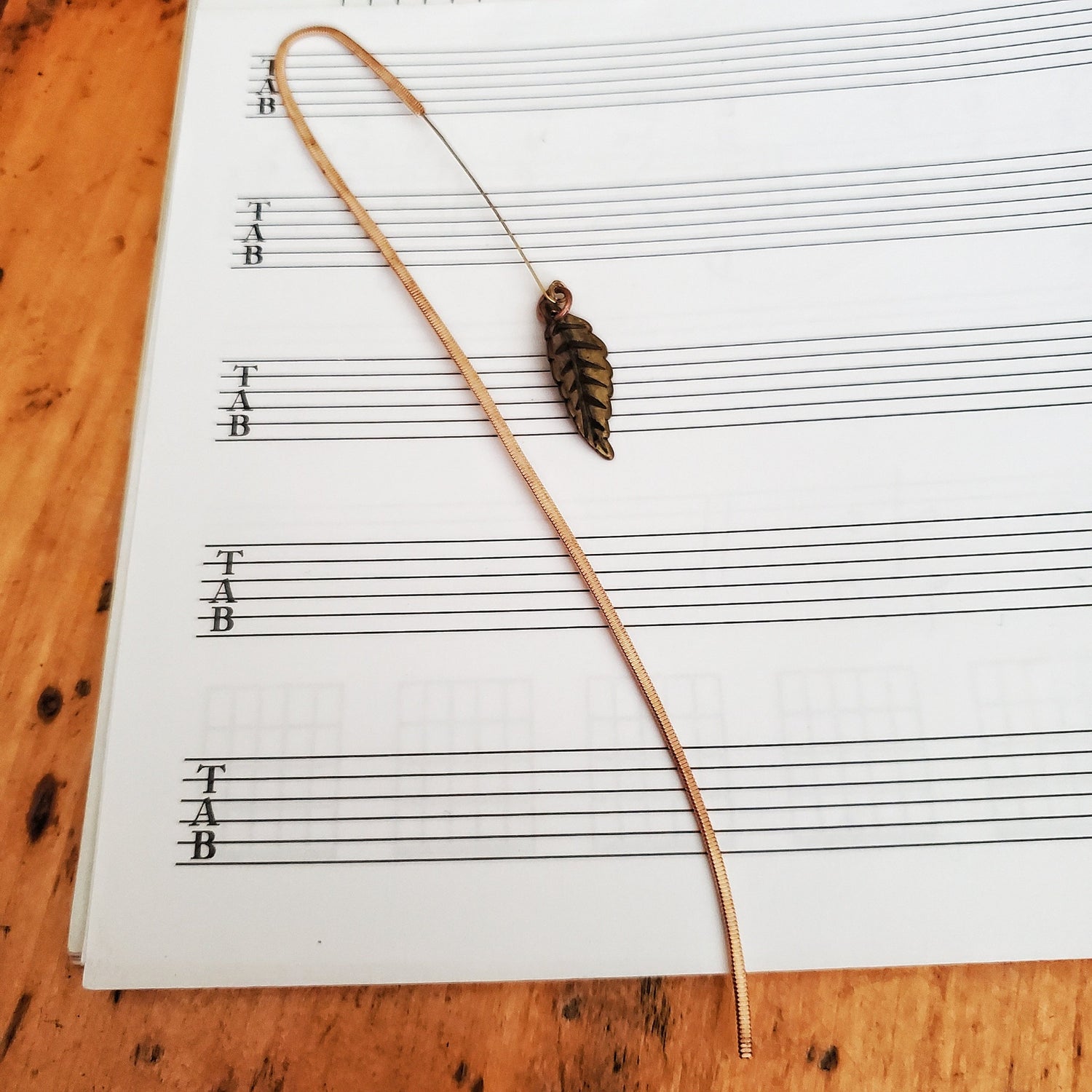 hook style bookmark made from an upcycled guitar string and a leaf shaped charm on blank sheet music