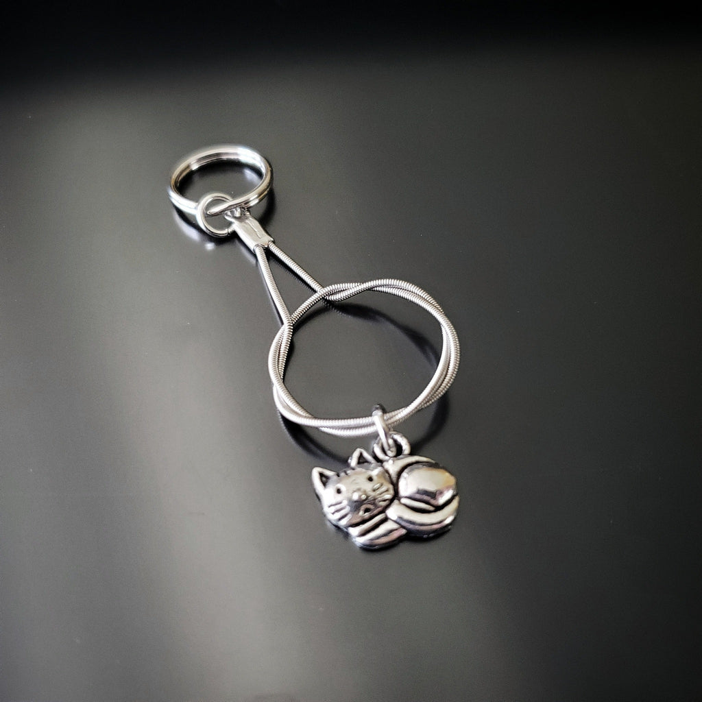 a silver coloured keychain made from an upcycled guitar string on which hangs a silver cat pendant - black background with grey shadow