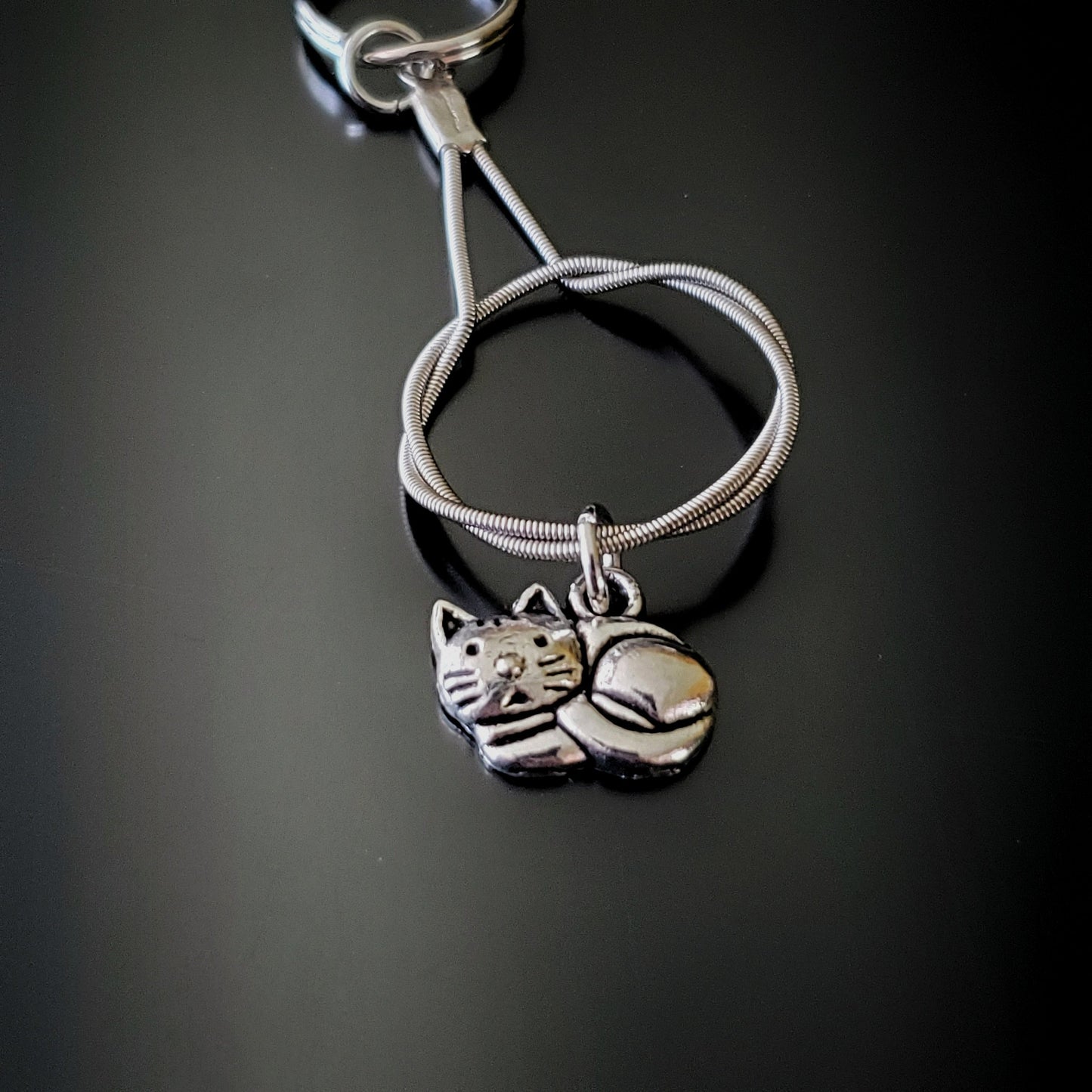 a silver coloured keychain made from an upcycled guitar string on which hangs a silver cat pendant - black background with grey shadow
