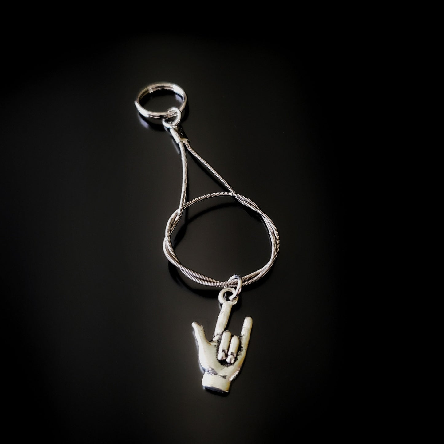 silver coloured keychain made from an upcycled guitar string on it is hanging a silver coloured pendant in the shape of a hand with the thumb, index and pinky fingers extended 
