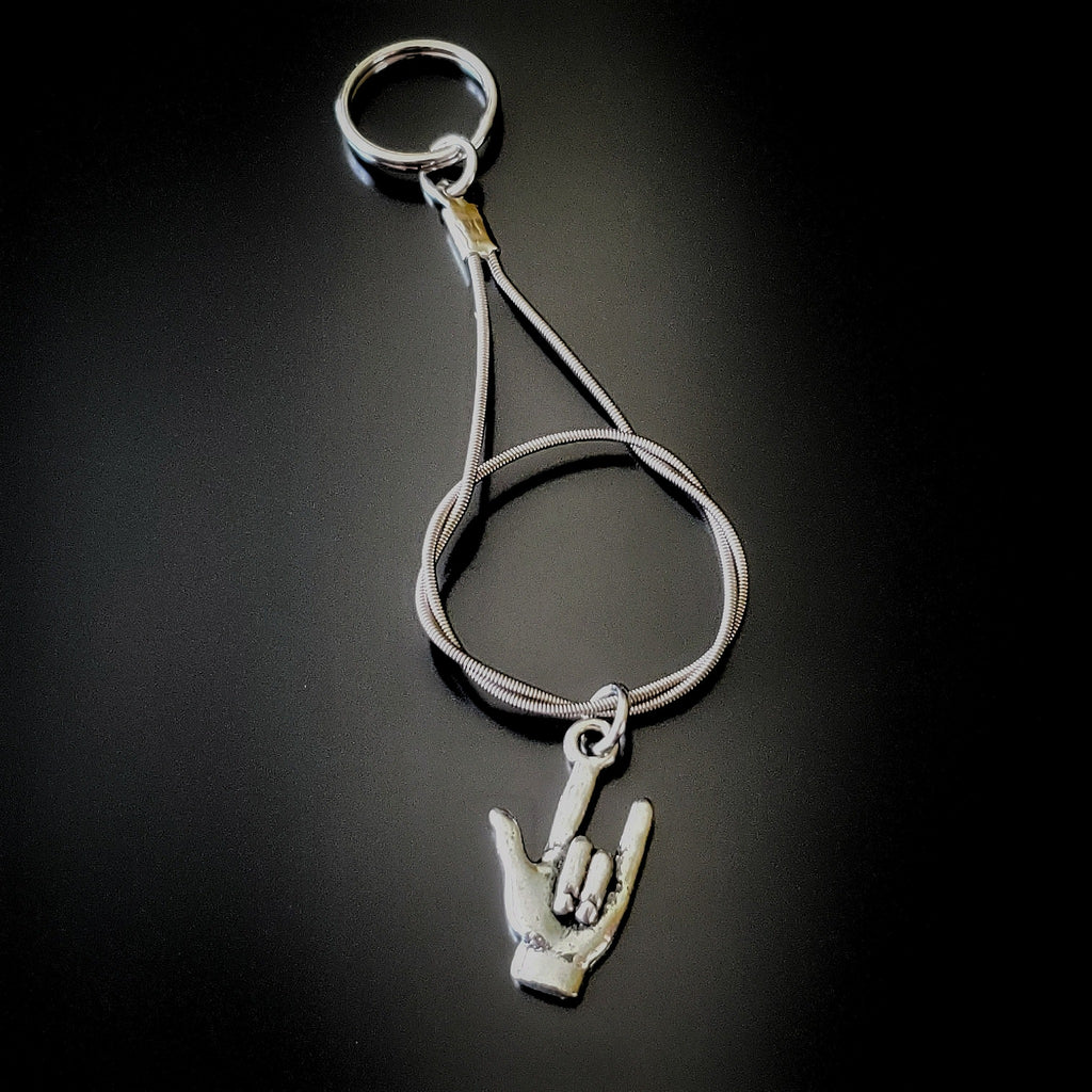 silver coloured keychain made from an upcycled guitar string on it is hanging a silver coloured pendant in the shape of a hand with the thumb, index and pinky fingers extended