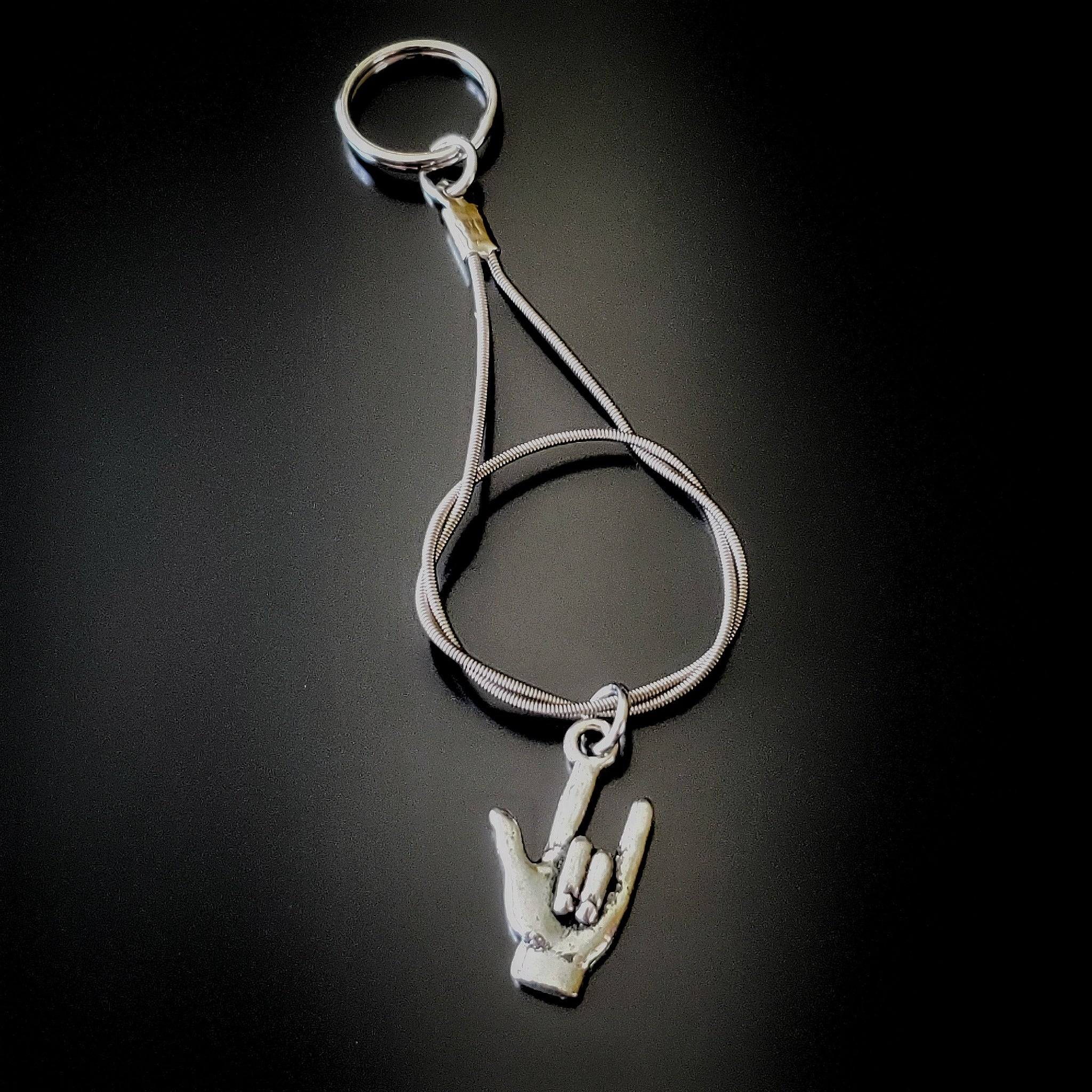 silver coloured keychain made from an upcycled guitar string on it is hanging a silver coloured pendant in the shape of a hand with the thumb, index and pinky fingers extended