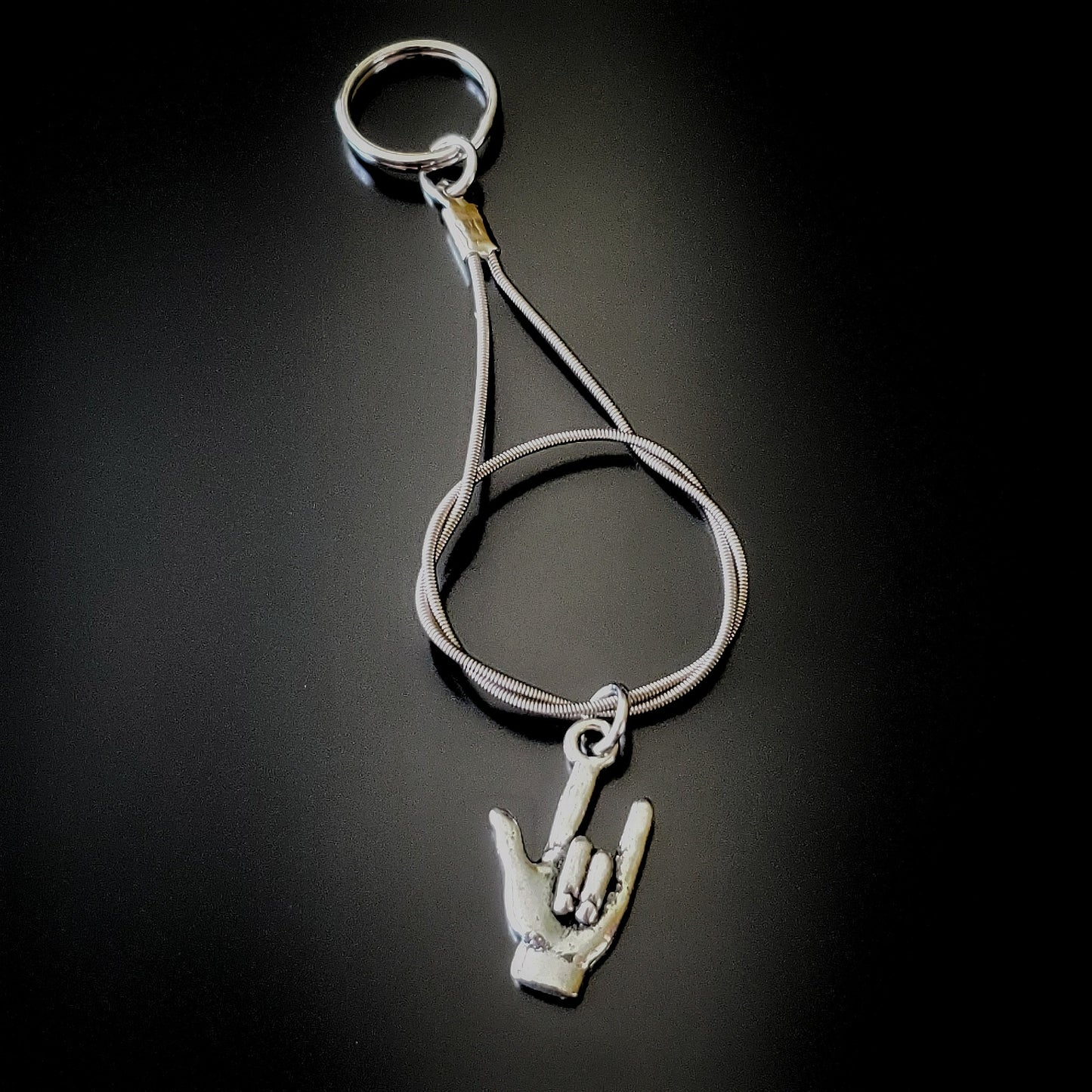 silver coloured keychain made from an upcycled guitar string on it is hanging a silver coloured pendant in the shape of a hand with the thumb, index and pinky fingers extended