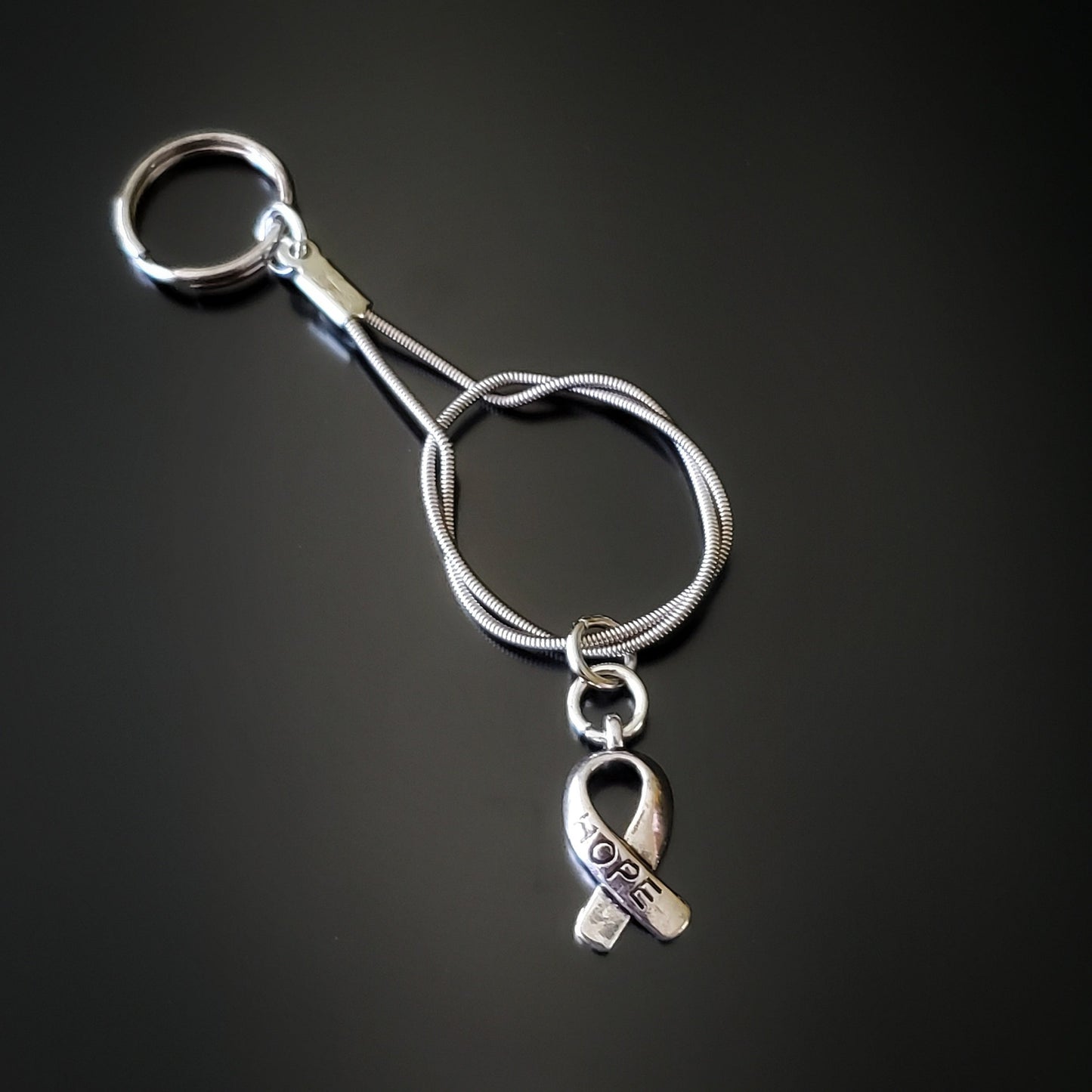 silver coloured keychain made from an upcycled guitar string with a silver coloured ribbon shaped charm with the word HOPE etched on it