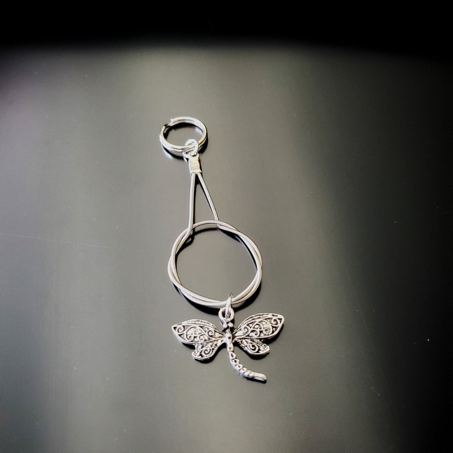 silver coloured keychain made from an upcycled guitar string on which hangs a silver coloured dragonfly pendant black and grey background