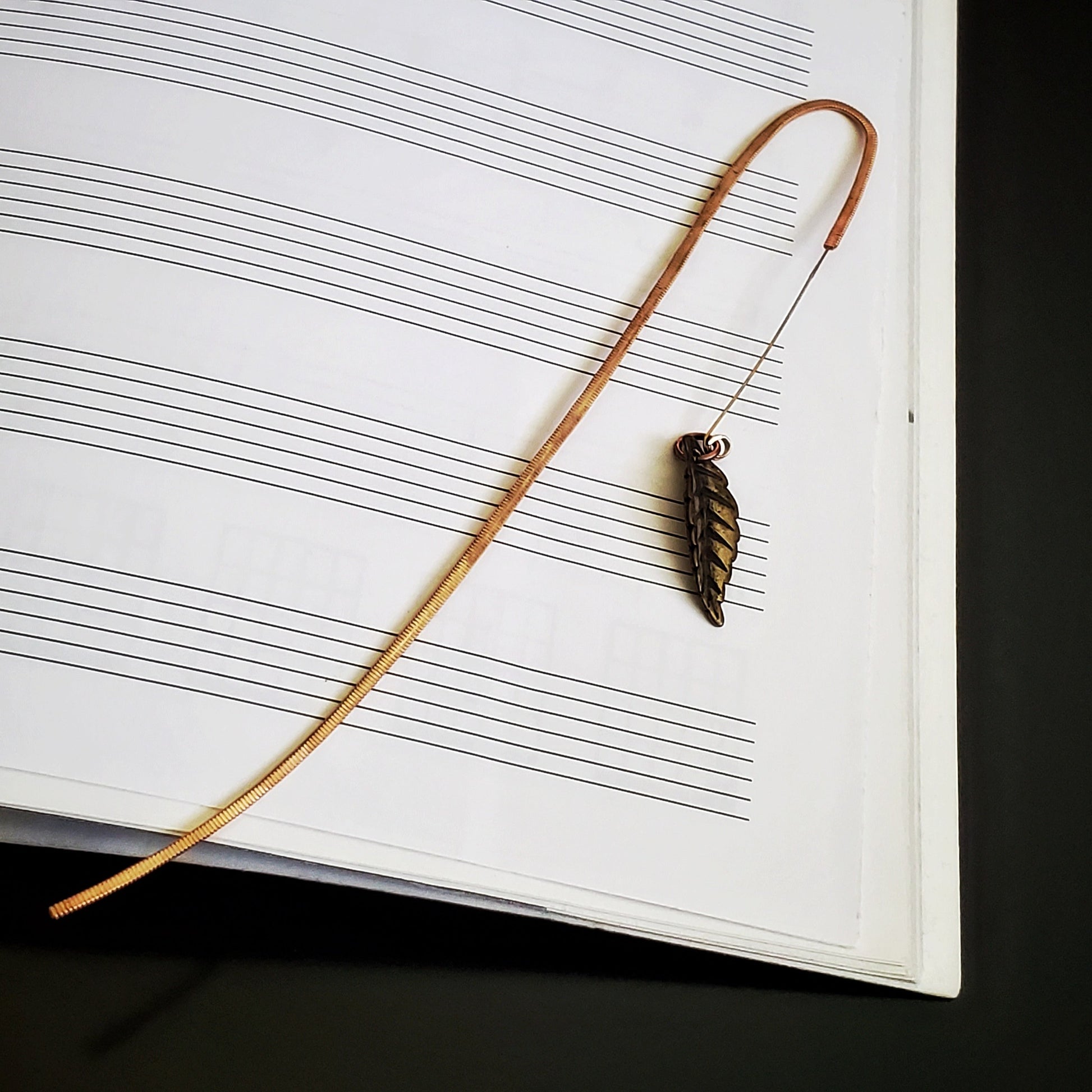 hook style bookmark made from an upcycled guitar string and a leaf shaped charm - background is blank sheet music