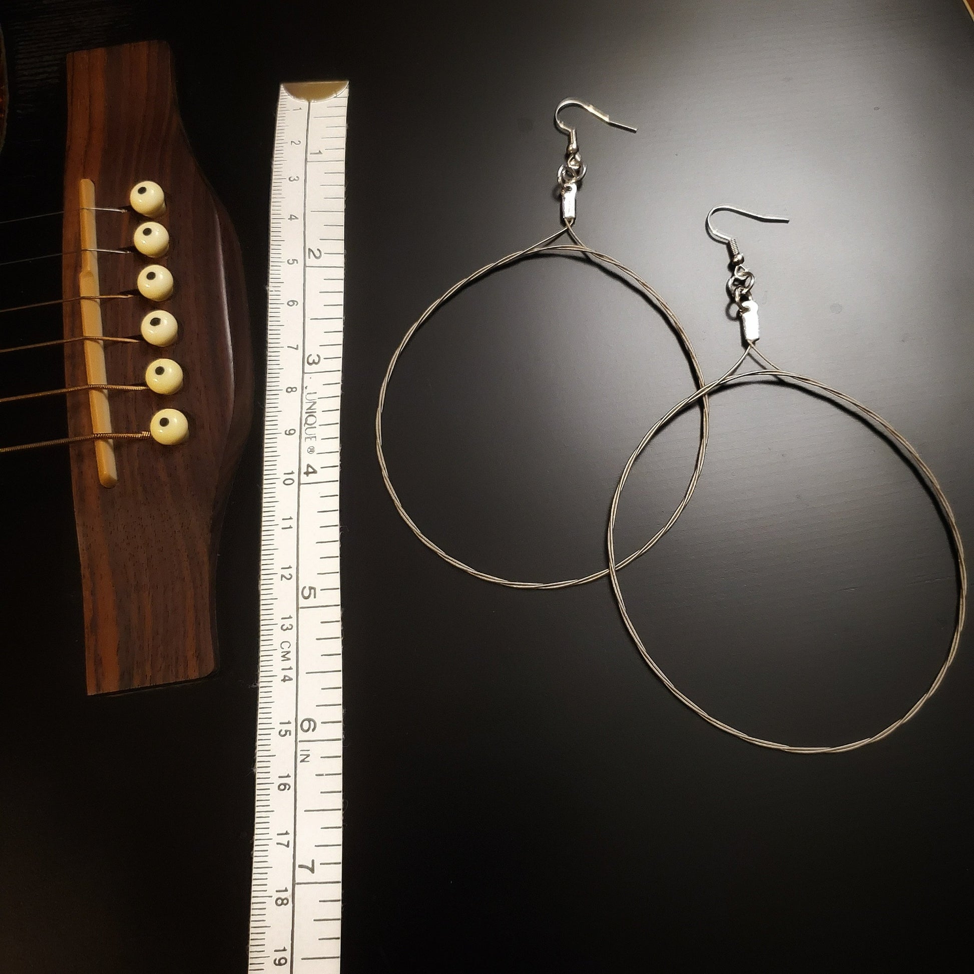pair of large hoop style earrings made from upcycled guitar strings on the left a white tape measure on the left of which is the bridge of a black guitar