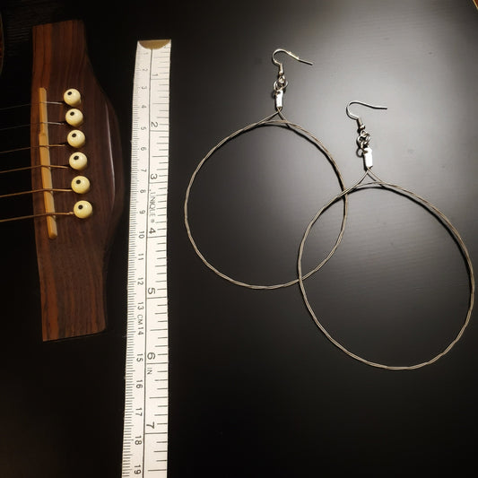 pair of large hoop style earrings made from upcycled guitar strings on the left a white tape measure on the left of which is the bridge of a black guitar