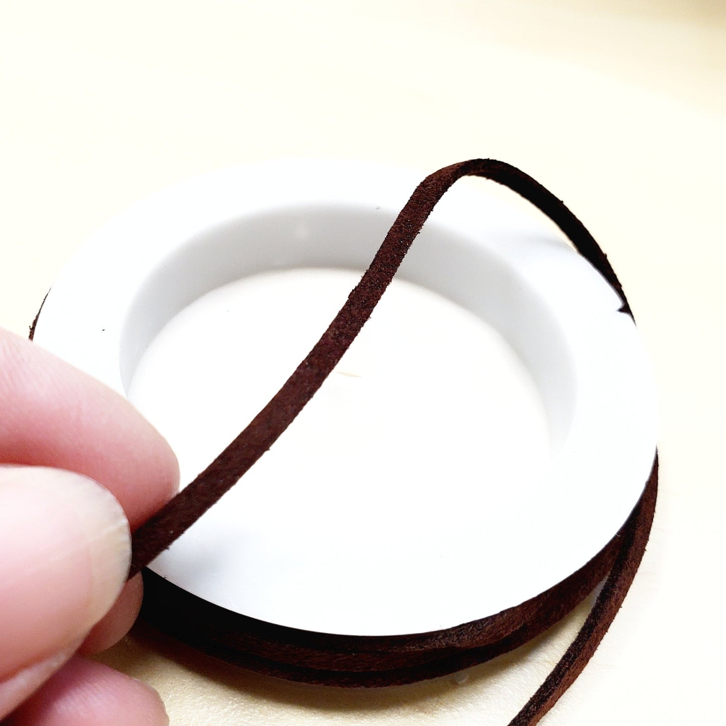 fingers holding a string of dark brown faux-suede coming from a white bobbin