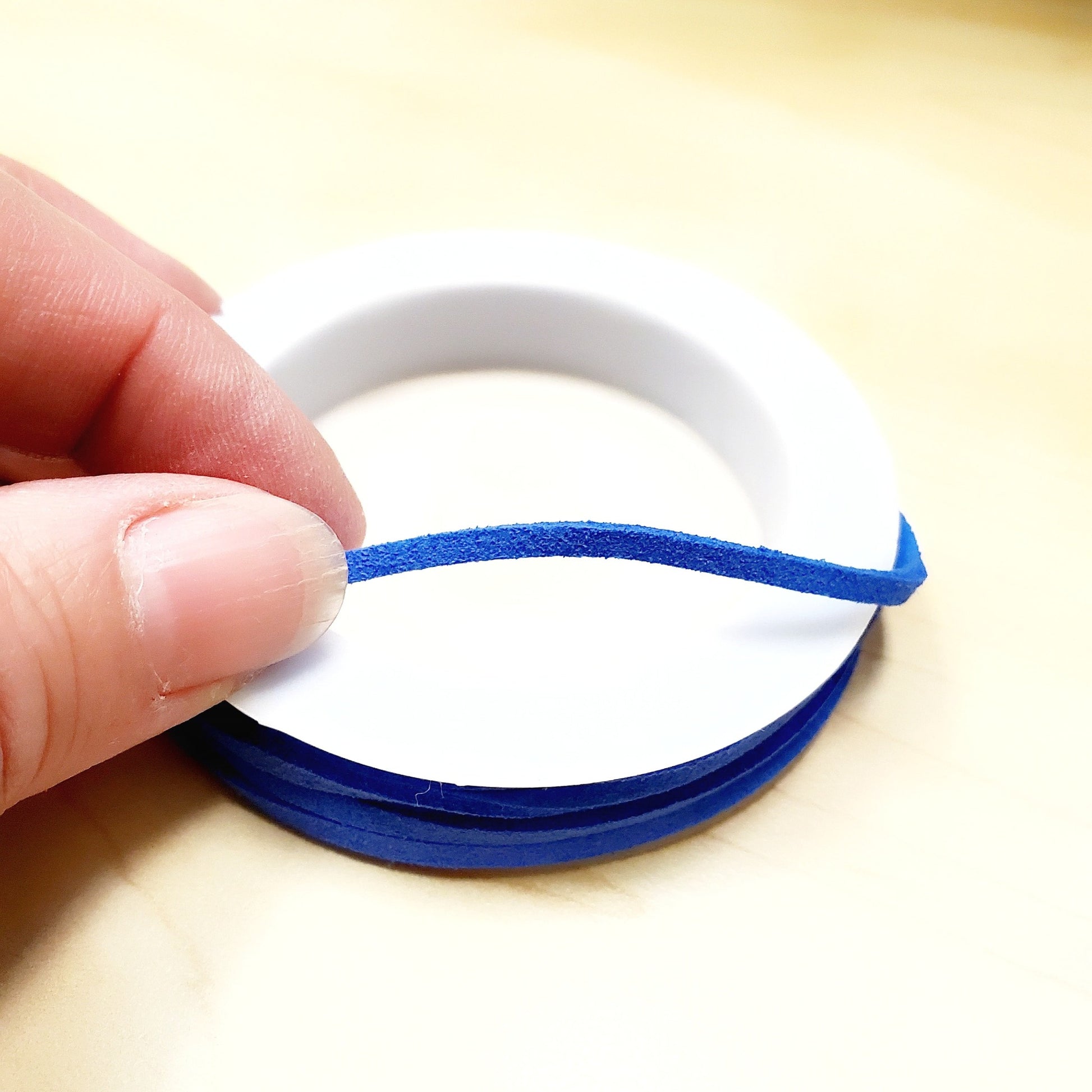 fingers holding a string of blue faux-suede coming from a white bobbin