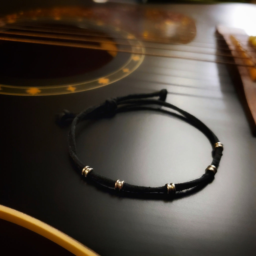 black bracelet made from a suede cord and silver coloured guitar string ballends sitting on the body of a black guitar