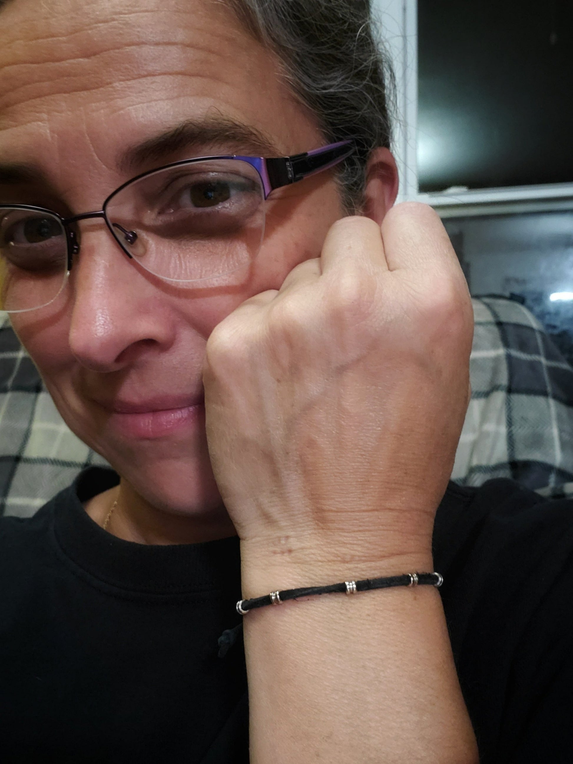 woman's head wearing purple rimed glasses her hand is in a fist next to her face - on her wrist is a black bracelet made from a suede cord and silver coloured guitar string ballends 