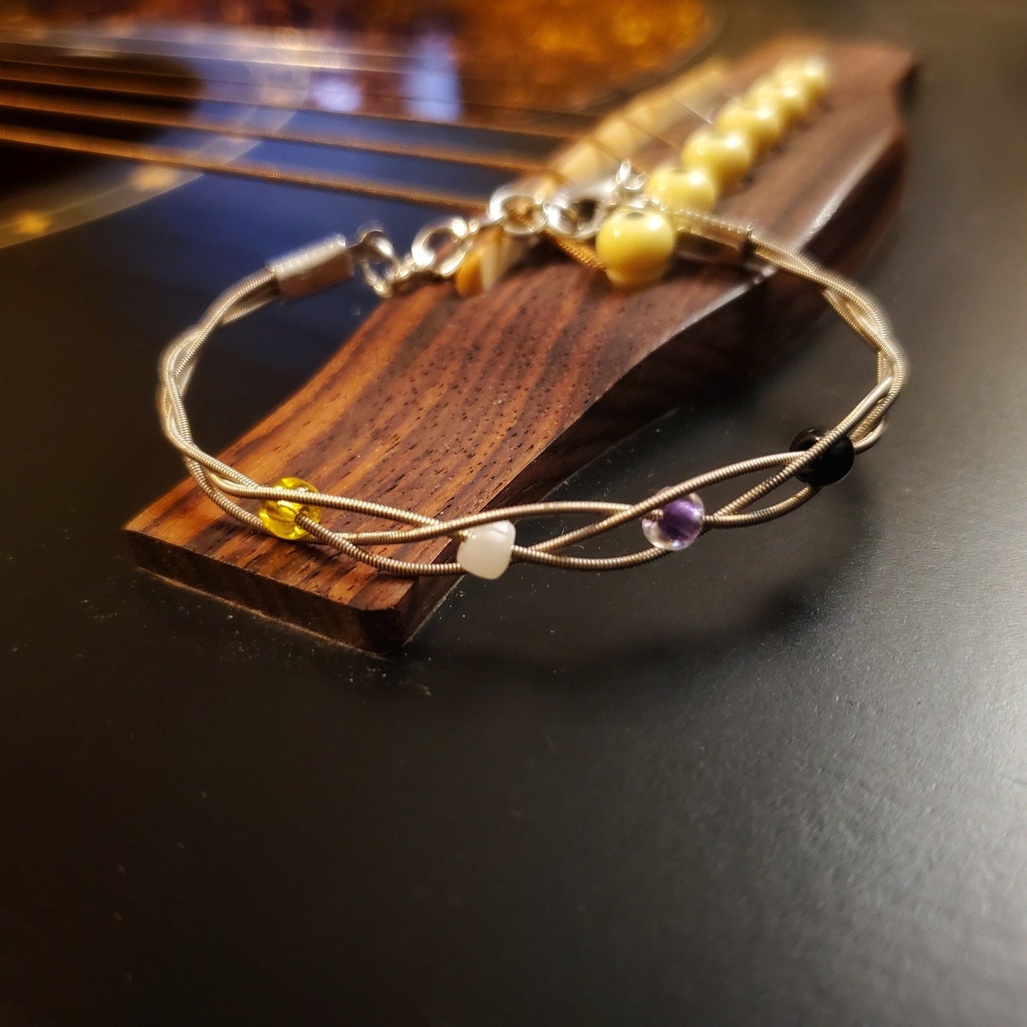clasp style bracelet made from braided upcycled guitar strings with glass beads representing the Non-Binary Pride flag sitting on the bridge of a black guitar