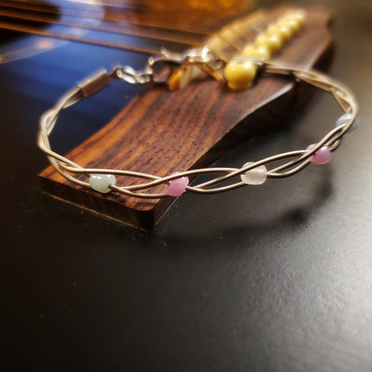 clasp style bracelet made from braided upcycled guitar strings with glass beads representing the Trans Pride flag sitting on the bridge of a black guitar