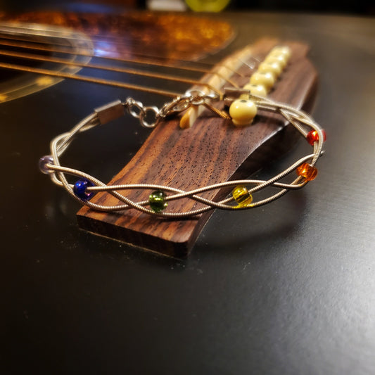 clasp style bracelet made from braided upcycled guitar strings with glass beads representing the LGBTQ Pride flag sitting on the bridge of a black guitar